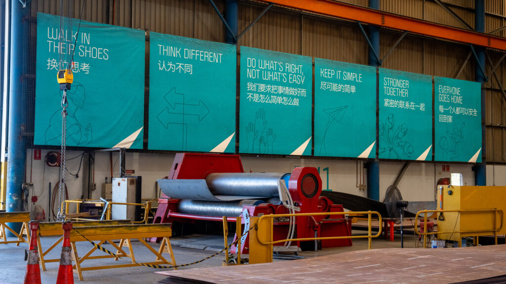 Photo des panneaux multilingues chez Schlam Forrestfield. Les panneaux indiquent : SE METTRE À LEUR PLACE, PENSER DIFFÉREMMENT, FAIRE CE QUI EST JUSTE PAS CE QUI EST FACILE, GARDER LA SIMPLICITÉ, PLUS FORTS ENSEMBLE, TOUT LE MONDE RENTRE CHEZ SOI. Un rouleau de tôle se trouve devant les panneaux.